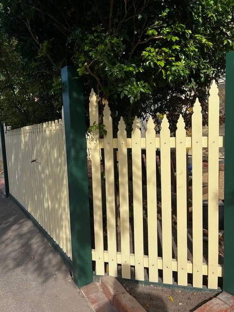 White picket fence gate with green post attached to a corrugated metal fence, surrounded by lush green foliage overhead