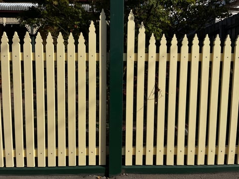 Cream-colored picket fence gates with pointed tops and a dark green center post, flanked by trees in background
