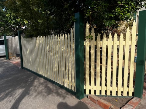 Green and white wooden picket fence gate with vertical slats on a brick pathway, surrounded by trees