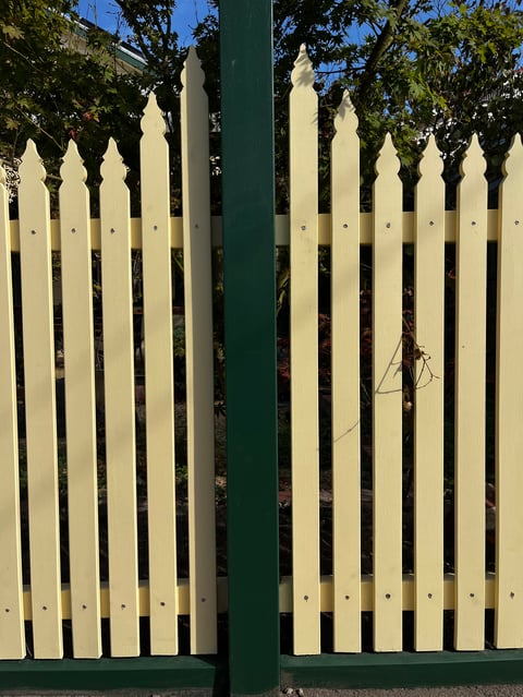 White picket fence gate with pointed posts on green frame, flanked by green trees under blue sky
