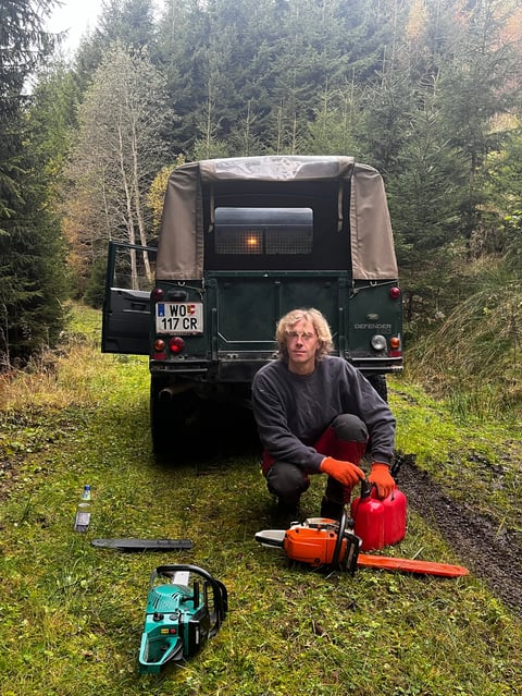 Man crouching in front of vintage Land Rover truck displaying chainsaws and tools in a forested setting
