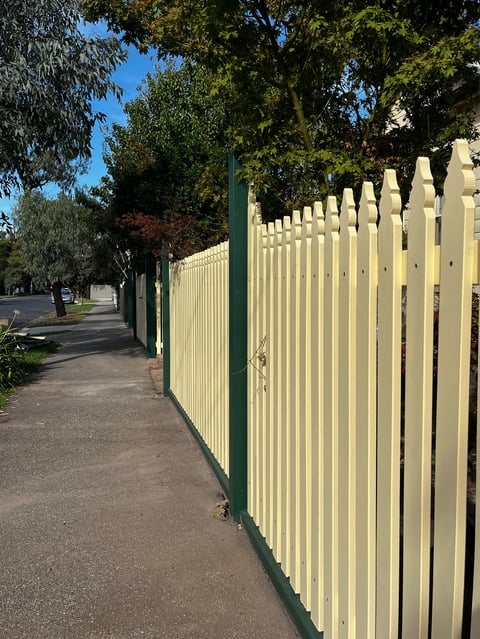 Cream and green picket fence lining a tree-lined residential street on a sunny day