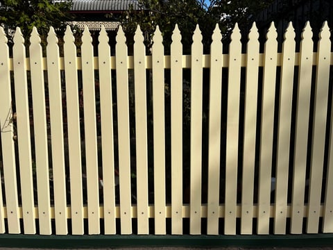 A row of tall, pointed white picket fence panels with trees visible in the background