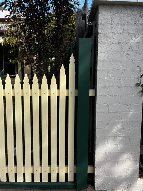 White wooden picket fence with green gate post next to a white brick wall and climbing vines