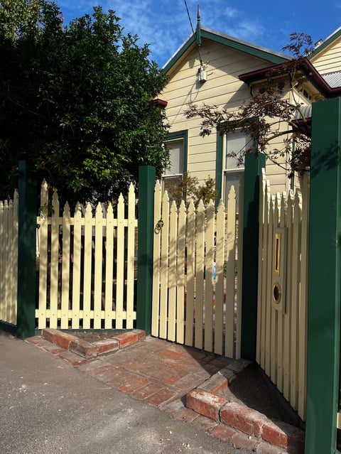 Cream and green cottage with white picket fence gate and brick pathway on sunny day