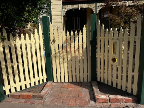 White picket gate with green posts open on brick pathway leading to Victorian house entrance