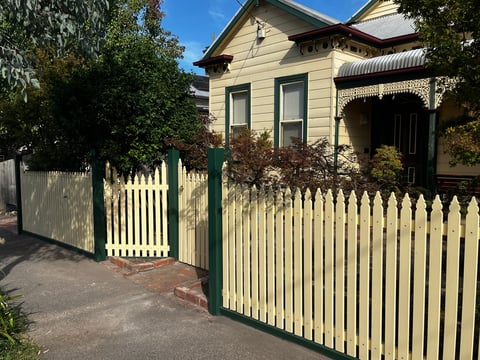 Historic cream and green cottage with white picket fence and gated entrance surrounded by green foliage